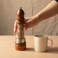 woodnestcraft wooden salt and pepper grinder with clear acrylic body held by hand next to a ceramic mug on a table